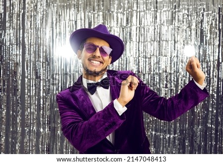 Happy funny young black man wearing tacky unfashionable purple velvet suit, hat and sunglasses dancing at party. Goofy ethnic guy dancing on concert show stage with shiny foil fringe studio background