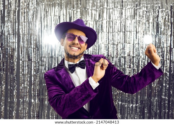 Happy funny young black man wearing tacky\
unfashionable purple velvet suit, hat and sunglasses dancing at\
party. Goofy ethnic guy dancing on concert show stage with shiny\
foil fringe studio\
background