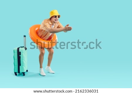 Happy funny young man wearing holiday shirt, summer hat, sun glasses and swim ring standing with suitcase on blue advertising copy space background and getting ready to jump in sea waters