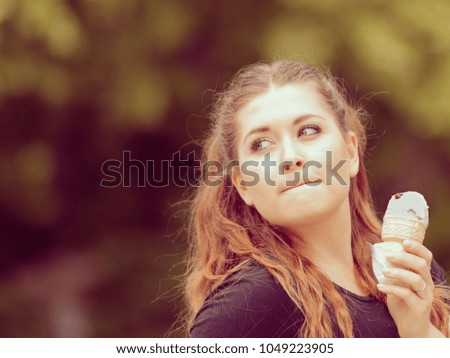 Happy funny young woman with long brown hair eating ice cream having fun enjoying her dessert during beautiful summer weather against green park