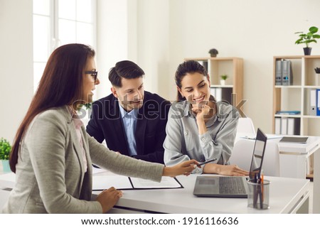 Happy future house buyers meeting real estate agent. Professional realtor talking to clients and offering flats options on computer. Smiling couple consulting bank worker or loan broker at her office