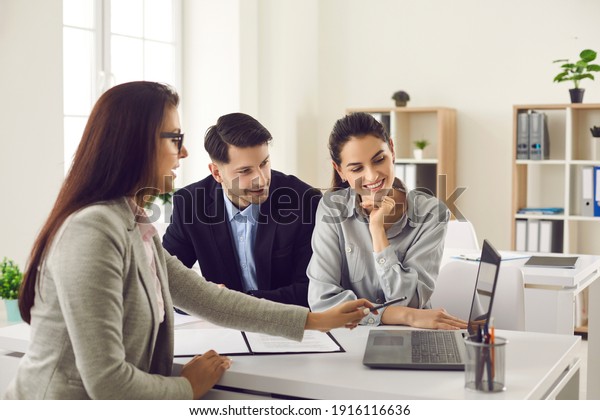 Happy future house buyers meeting real estate
agent. Professional realtor talking to clients and offering flats
options on computer. Smiling couple consulting bank worker or loan
broker at her office