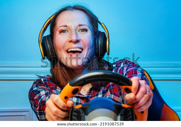 Happy gamer woman in\
headphones with hands on console steering wheel of a laughing\
playing race game
