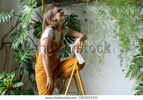Happy gardener woman holding pulverizer spray, smiling,\
takes a break from work, sitting on stepladder, wearing glasses and\
orange overalls. Greenery at home. Love of plants. Indoor cozy\
garden. 