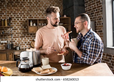 Happy Gay Couple Eating Toasts With Jam In Kitchen At Morning