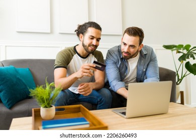 Happy Gay Couple Enjoying Online Shopping Together On The Laptop And Paying With A Credit Card While Relaxing On The Sofa