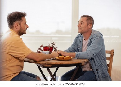 Happy gay couple holding hands and sharing a tender moment while having breakfast together at their dining table, enjoying muffins and coffee in a bright and cozy home environment - Powered by Shutterstock