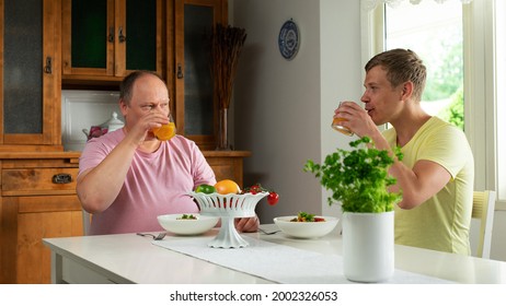 A Happy Gay Couple Looking At Each Other While Drinking Orange Juice And Eating Pasta From White Bowls Behind A Big White Table That Has A Fruit Bowl And A Vase Of Flowers.