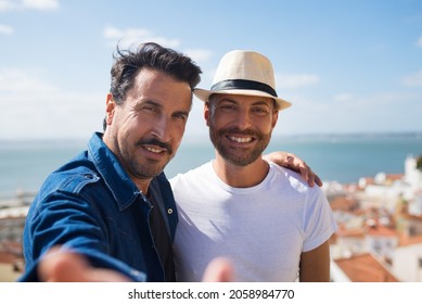 Happy gay couple on sunny day. Two men in casual clothes, standing on balcony, looking at camera, sea in background. Love, affection, relationship concept - Powered by Shutterstock