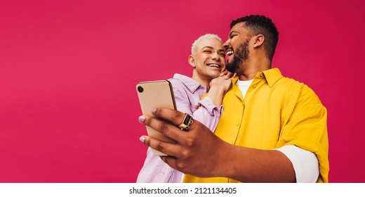 Happy gay couple taking a selfie while standing against a pink background. Cute gay couple smiling cheerfully while using a smartphone. Young gay couple creating memories together in a studio.
