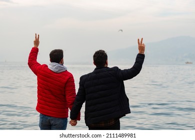 Happy gay couple watching sunset on the coast. Back view. They are holding hands and making peace signs. LGBT, freedom, understanding in tolerance, romance concept. Horizontal banner website. 