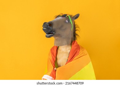 Happy gay female in surreal horse head mask wrapped with LGBT rainbow flag isolated on orange background