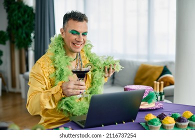 Happy gay man wearing carnival costume and toasting to someone during video call over laptop while celebrating Mardi Gras at home.