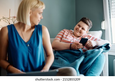 Happy Gay Woman Texting On Cell Phone And Communicating With Her Girlfriend Who Is Using Laptop At Home. 