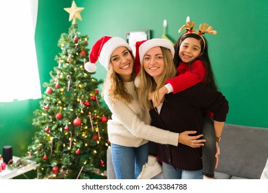 Happy Gay Women And Moms With A Santa Hat Piggybacking Her Cute Daughter. Multiracial Family Hugging During The Christmas Holidays 