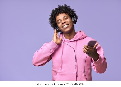 Happy gen z African American guy wearing headphones holding mobile using cellphone dancing enjoying listening online streaming music on cell phone, standing isolated on light purple background.