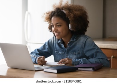 Happy gen Z Black teenage student girl chatting online with teacher, using headphones and laptop at home, writing notes, preparing for school test, college exam, watching learning webinar