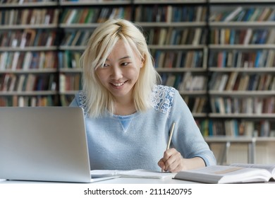 Happy gen Z blonde Asian student girl doing homework in college library, enjoying work on study project, research article, writing essay draft, consulting tutor on video call, watching webinar - Powered by Shutterstock