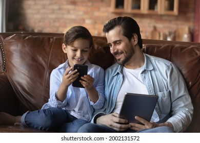 Happy Gen Z Boy Kid And Daddy Using Gadgets At Home, Playing, Compete In Online Game, Holding Digital Tablet And Mobile Phone, Browsing Internet, Using Learning App, Making Video Call. Family Activity