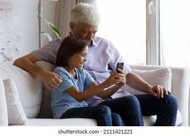 Happy Gen Z Grandson Boy And Senior Gray Haired Grandpa Using Smartphone At Home Together, Sitting On Couch, Making Video Call, Taking Selfie, Enjoying Leisure, Talking To Family On Mobile Phone