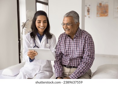 Happy geriatric young doctor and senior Indian patient man discussing modern technology, healthcare, using digital tablet together, looking at display in practitioner office, laughing - Powered by Shutterstock