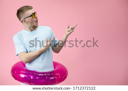 happy gesticulating young businessman wearing white T-shirt in inflatable swim ring calling for vacation in sunglasses isolated on pink background copy space. holiday concept