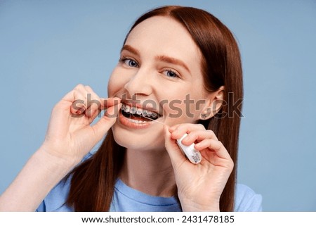 A happy, ginger haired woman with dental brackets, meticulously cleaning her teeth with floss, isolated on a blue background. Highlights tooth care concept