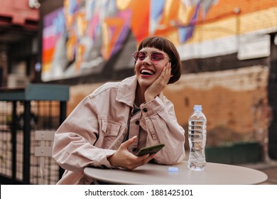 Happy girl in beige denim jacket smiles sincerely outside. Short-haired teen in bright glasses and light clothes holding phone and laughing at street..