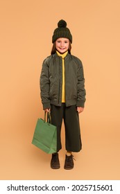 Happy Girl In Bomber Jacket And Brown Hat Holding Shopping Bag On Beige