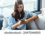 Happy girl checking social media while holding smartphone at home. Smiling young brunette woman using mobile phone app to play games, shop online, order delivery while relaxing on sofa. Scrolling.