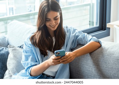 Happy girl checking social media while holding smartphone at home. Smiling young brunette woman using mobile phone app to play games, shop online, order delivery while relaxing on sofa. Scrolling. - Powered by Shutterstock