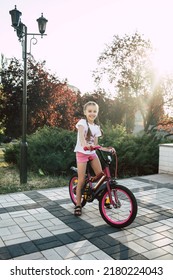 Happy Girl Child Rides A Red Bike In The Park During Sunset. Cute Little Girl Is Dressed In Summer Clothes And Smiling. The Child Loves To Ride A Bike. Happy Childhood And Children's Love For Cycling