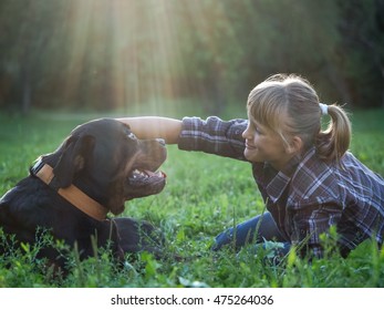 Happy Girl And Dog In The Sunshine. The Dog Is Big And Scary - Rottweiler. Green Grass, Park, Sunset