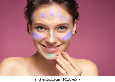 Happy girl doing makeup using concealer. Photo of beautiful brunette girl on pink background. Skin care concept