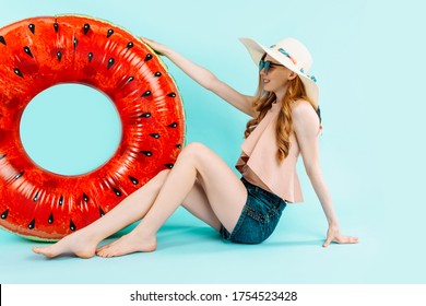 happy girl, dressed in a swimsuit and a summer hat, sits with an inflatable swimming circle, resting isolated on a blue background