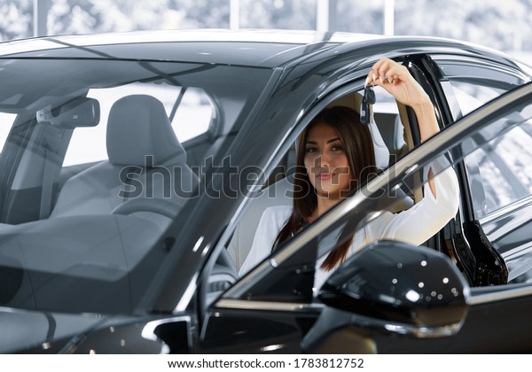 A happy girl driver
smiles while holding the keys to her new car. The girl is sitting
in a new car at the dealership, showing the car keys. Ownership,
driving concept