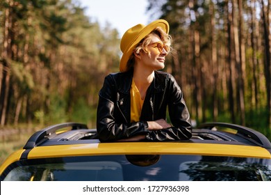 Happy Girl Enjoy Summer Sunlight At Yellow Car Sunroof, Road Trip Joy. Dreamy Woman In Jacket, Hat And Sunglasses Leaning On Auto Moon Roof At Forest. Trendy Blonde Weekend Adventure Lifestyle