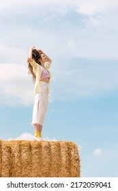 Happy girl enjoys fresh air in the countryside. Young beautiful woman with long hair stands on haystack against blue sky