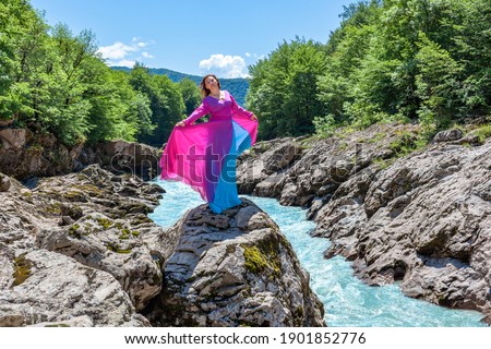 A happy girl in a flowing dress stands on the edge of a canyon above a stormy mountain river on a summer sunny day.