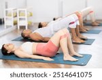 Happy girl and group of women and men lying on mats and doing stretch exercises with resistance bands in light fitness room