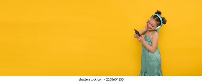 Happy Girl In Headphones Holds A Mobile Phone And Listens To Music, Not Looking At The Camera, Banner, Yellow Background. High Quality Photo