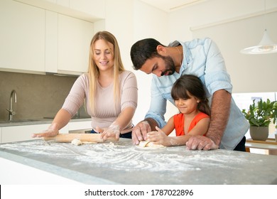 Happy Girl And Her Dad Helping Mom To Knead Dough On Kitchen Counter With Flour Messy. Young Couple And Their Daughter Baking Buns Or Pies Together. Family Cooking Concept