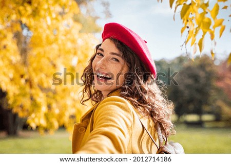 Happy girl holding hand and looking behind while walking in the park. Beautiful woman relaxing at the park wearing winter clothing. Young smiling woman pulling her boyfriend and looking at camera.