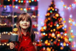
Happy Girl Holding A Microphone Singing Carols. Cheerful Child Performing Christmas Songs For Her Family At Home
