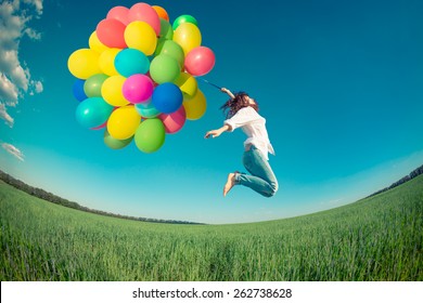 Happy girl jumping with colorful toy balloons outdoors. Young woman having fun in green spring field against blue sky background. Freedom concept