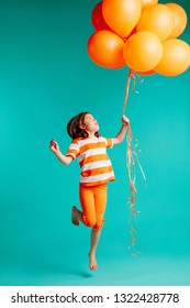 Happy girl jumping with orange balloons in hand. Full length of cute girl child with bunch of helium balloons on blue background.