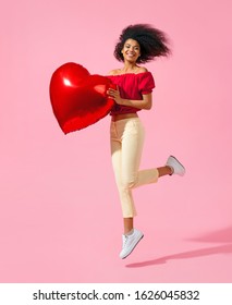 Happy girl jumping with red heart shape balloon. Photo of smiling young girl in love on pink background. Valentine's Day