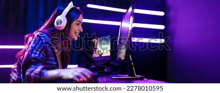 Happy girl live streaming a video game, sitting in front of a multi monitor with a microphone for commentary and a headset for communication. Female engaging with fellow gamers on an online platform.