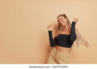 Happy girl with long dreadlocks dressed in black long sleeve top dancing with hands up on light brown backdrop, happiness concept, copy space