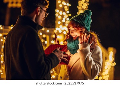 A happy girl is looking at christmas surprise in a box while her boyfriend is showing box to her on city street with christmas decorations. A festive couple exchanging christmas gifts on christmas. - Powered by Shutterstock